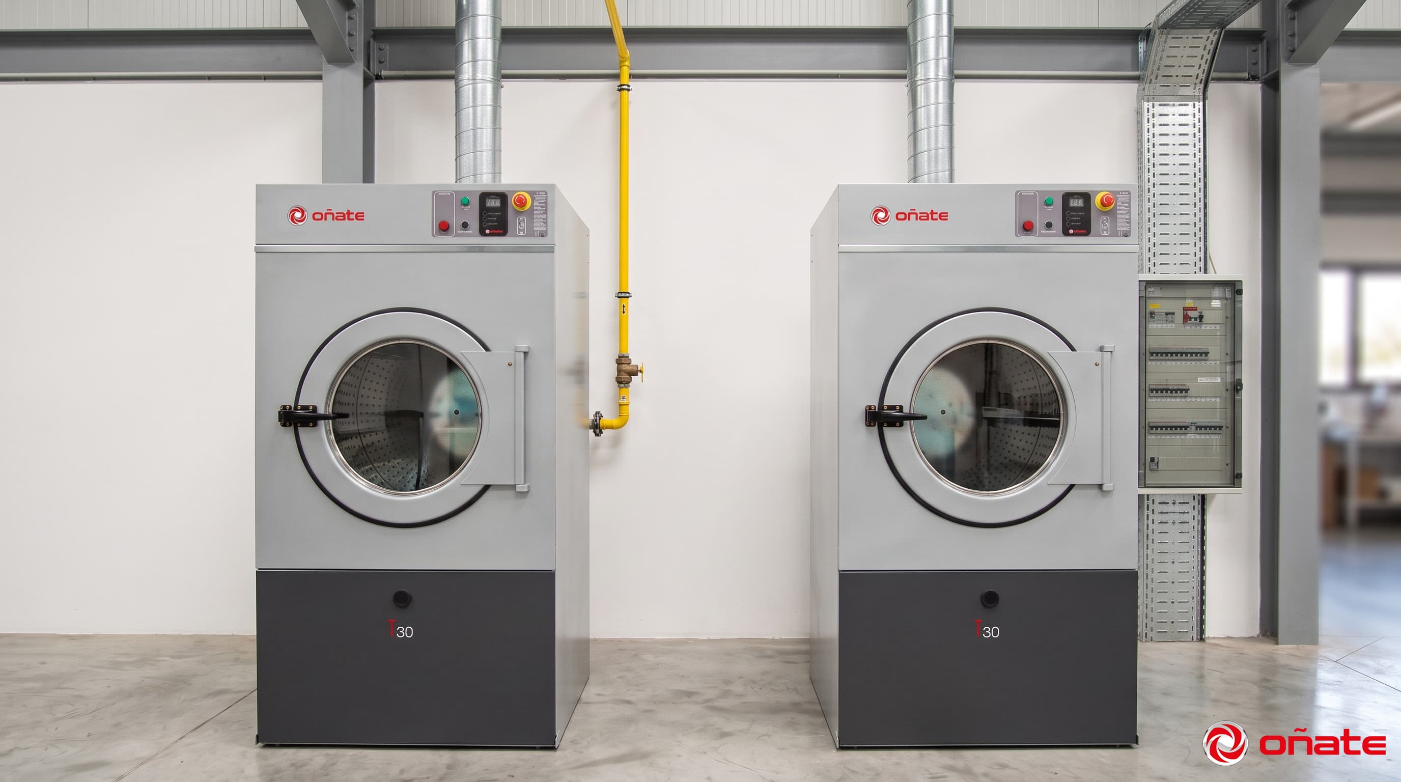 Dos secadoras industriales Oñate T30 idénticas lado a lado: la de la izquierda alimentada por un caño de gas amarillo que baja del techo, la de la derecha por una bandeja portacables galvanizada y un tablero eléctrico de pared